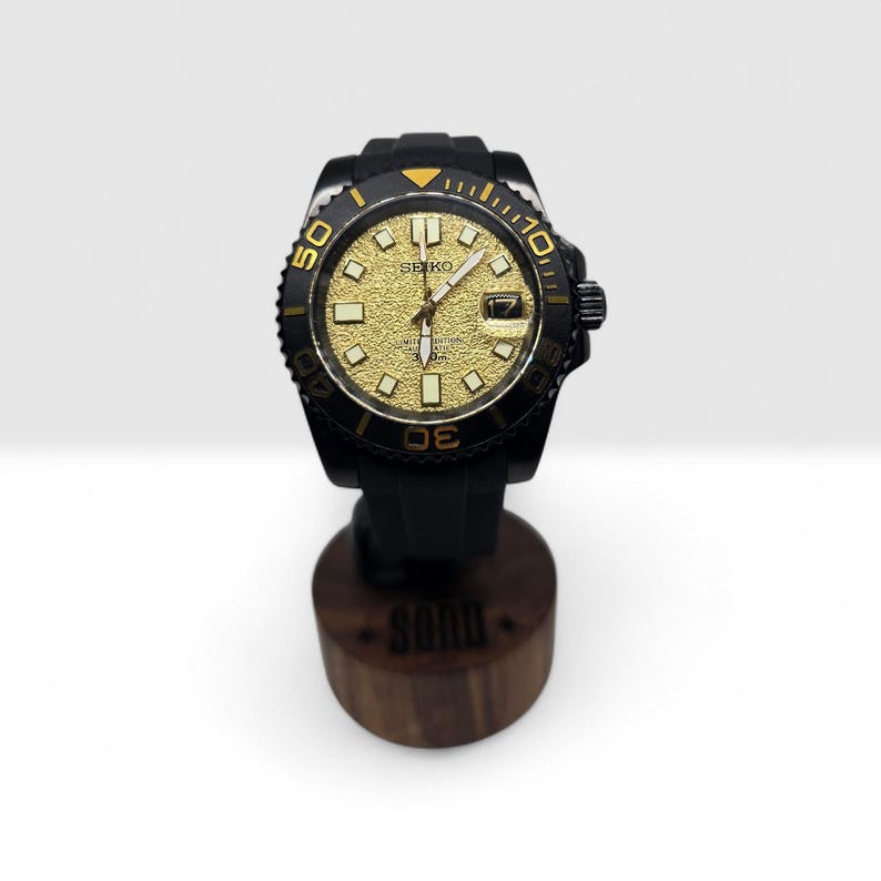 Pu&ograve; includere: Un orologio da polso Seiko con quadrante dorato testurizzato, indici luminosi, ghiera girevole nera con numeri dorati e cinturino in gomma nera. Il quadrante mostra il logo Seiko e una finestrella per la data in posizione a ore tre.