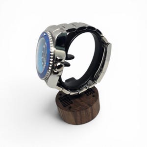 May include: A silver-tone wristwatch with a blue bezel and a black dial, featuring a metallic link bracelet. The watch is displayed on a cylindrical dark wood stand that has the word "SOND" engraved on its side with small star accents.