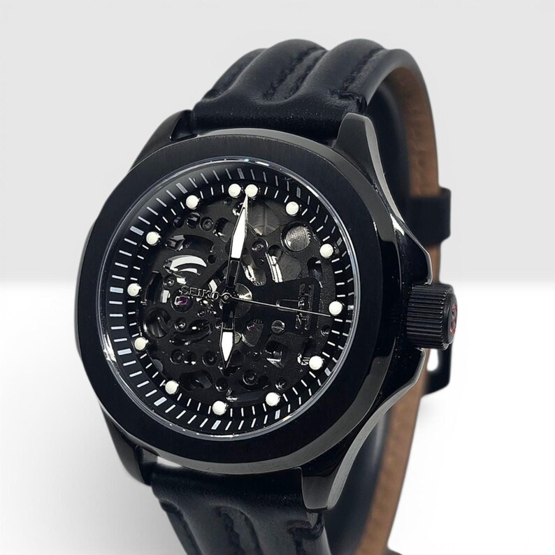 May include: A black Seiko wristwatch with a skeleton dial revealing the internal mechanical movement. The watch features a round black case, white hour markers, silver hands, and a black leather strap with prominent raised vertical stitching.