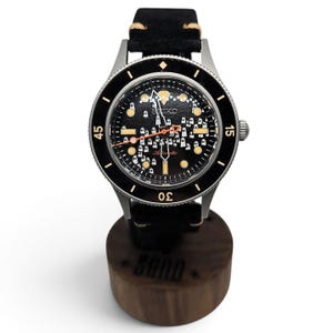 May include: An automatic wristwatch with a black dial featuring a repeating white ghost pattern, silver-tone hands, and a black rotating bezel with gold-tone markers. The watch has a black leather strap with tan stitching and a silver-tone case.