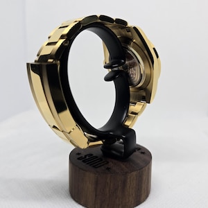 Op de afbeelding: Een goudkleurig polshorloge tentoongesteld op een zwarte standaard met een houten voet. Het horloge is zo geplaatst dat de band en wijzerplaat te zien zijn. De standaard is voorzien van het woord "SOND".