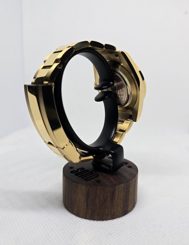 Op de afbeelding: Een goudkleurig polshorloge tentoongesteld op een zwarte standaard met een houten voet. Het horloge is zo geplaatst dat de band en wijzerplaat te zien zijn. De standaard is voorzien van het woord "SOND".