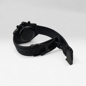 May include: A matte black wristwatch featuring a circular case and a textured silicone strap. The watch includes a black deployment clasp mechanism and a series of adjustment holes along the band, showcasing a monochromatic design throughout.