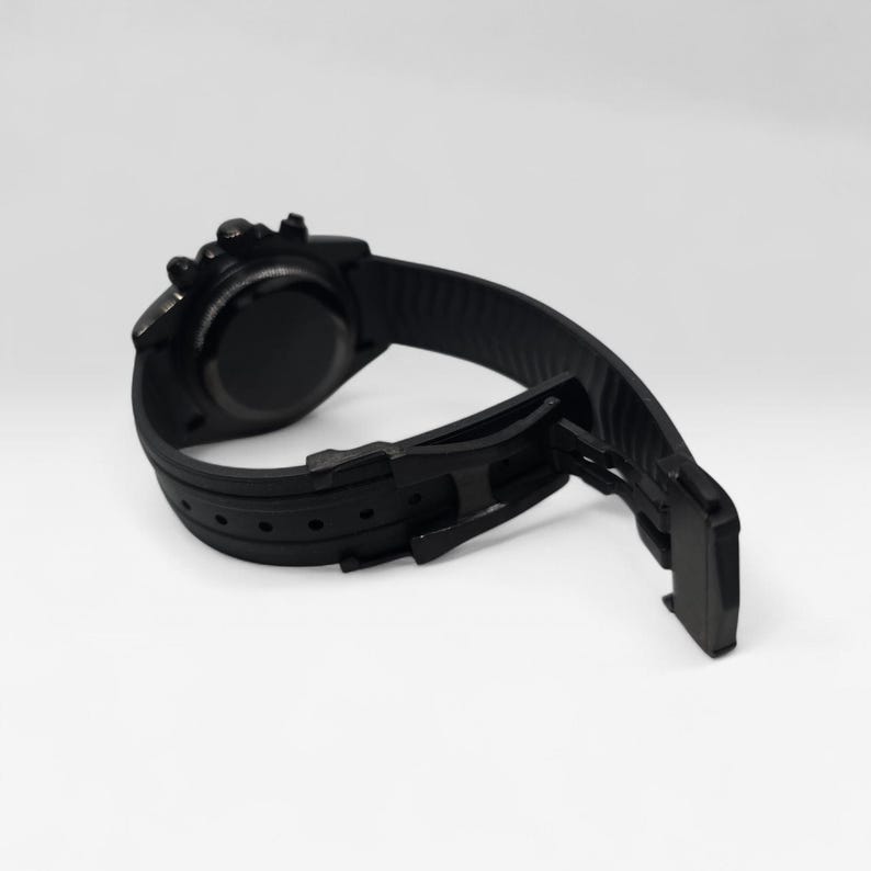 May include: A matte black wristwatch featuring a circular case and a textured silicone strap. The watch includes a black deployment clasp mechanism and a series of adjustment holes along the band, showcasing a monochromatic design throughout.