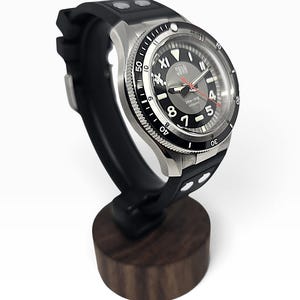 Puede incluir: Un reloj de pulsera autom&aacute;tico de acero inoxidable con esfera negra, n&uacute;meros blancos y segundero rojo. El reloj cuenta con una correa de caucho negra con remaches plateados y est&aacute; montado sobre un soporte de madera oscura.