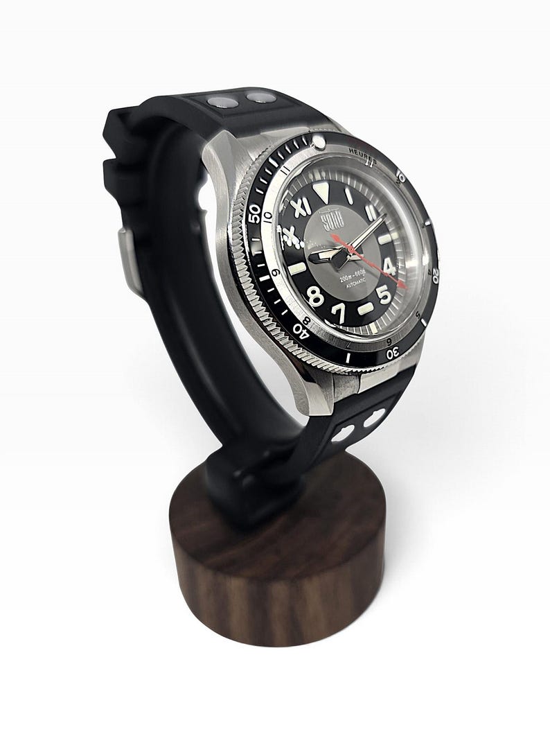 Puede incluir: Un reloj de pulsera autom&aacute;tico de acero inoxidable con esfera negra, n&uacute;meros blancos y segundero rojo. El reloj cuenta con una correa de caucho negra con remaches plateados y est&aacute; montado sobre un soporte de madera oscura.