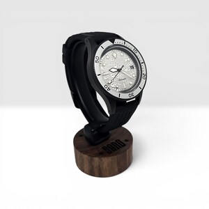 May include: An automatic wristwatch with a black case, a textured black rubber strap, and a silver-toned dial featuring a map pattern. The watch is mounted on a cylindrical wooden stand with the word SOND printed on the front surface.