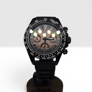 K&ouml;nnte beinhalten: Eine schwarze Chronographen-Armbanduhr mit dunkelbraunem Zifferblatt, drei Hilfszifferbl&auml;ttern, leuchtenden Zeigern und einer Tachymeterskala auf der L&uuml;nette. Die Uhr verf&uuml;gt &uuml;ber ein schwarzes Silikonarmband sowie eine Krone mit zwei Dr&uuml;ckern.