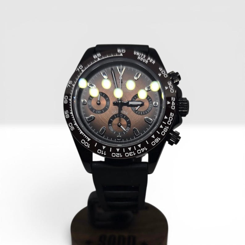 K&ouml;nnte beinhalten: Eine schwarze Chronographen-Armbanduhr mit dunkelbraunem Zifferblatt, drei Hilfszifferbl&auml;ttern, leuchtenden Zeigern und einer Tachymeterskala auf der L&uuml;nette. Die Uhr verf&uuml;gt &uuml;ber ein schwarzes Silikonarmband sowie eine Krone mit zwei Dr&uuml;ckern.