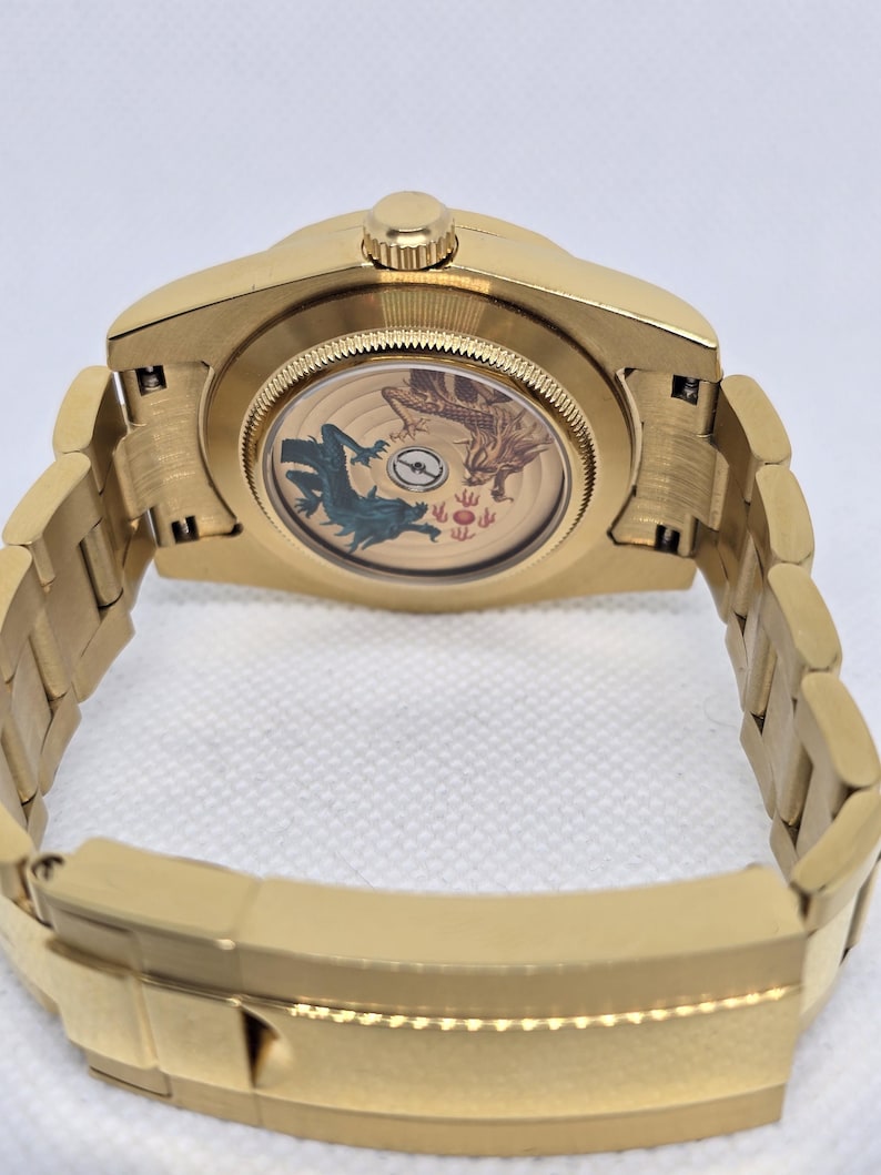 Op de afbeelding: Goudkleurig polshorloge met een gedetailleerde achterkant met een draak en andere symbolen. Het horloge heeft een gouden band en een kroon. De transparante achterkant onthult het interne mechanisme en het ontwerp.