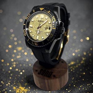 Pu&ograve; includere: Un orologio da polso Seiko con cassa nera e quadrante dorato testurizzato. L'orologio presenta una lunetta girevole nera con indicatori dorati, un cinturino in gomma nera e una finestrella per la data, esposto su un supporto in legno.