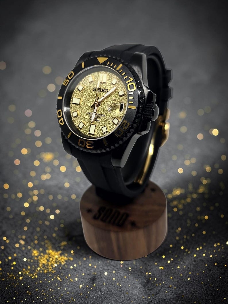 Pu&ograve; includere: Un orologio da polso Seiko con cassa nera e quadrante dorato testurizzato. L'orologio presenta una lunetta girevole nera con indicatori dorati, un cinturino in gomma nera e una finestrella per la data, esposto su un supporto in legno.