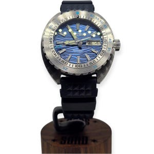 May include: A stainless steel wristwatch with a blue wave-patterned dial and a black rubber strap. The watch has a rotating bezel with minute markers and a date display. The watch is mounted on a wooden stand with the word "SUMO" engraved on it.