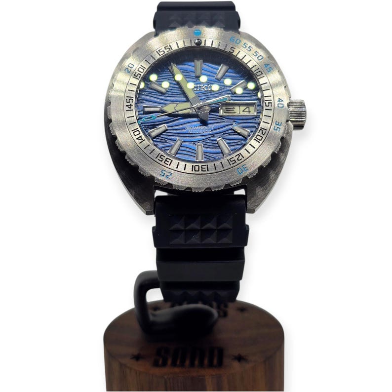 May include: A stainless steel wristwatch with a blue wave-patterned dial and a black rubber strap. The watch has a rotating bezel with minute markers and a date display. The watch is mounted on a wooden stand with the word "SUMO" engraved on it.