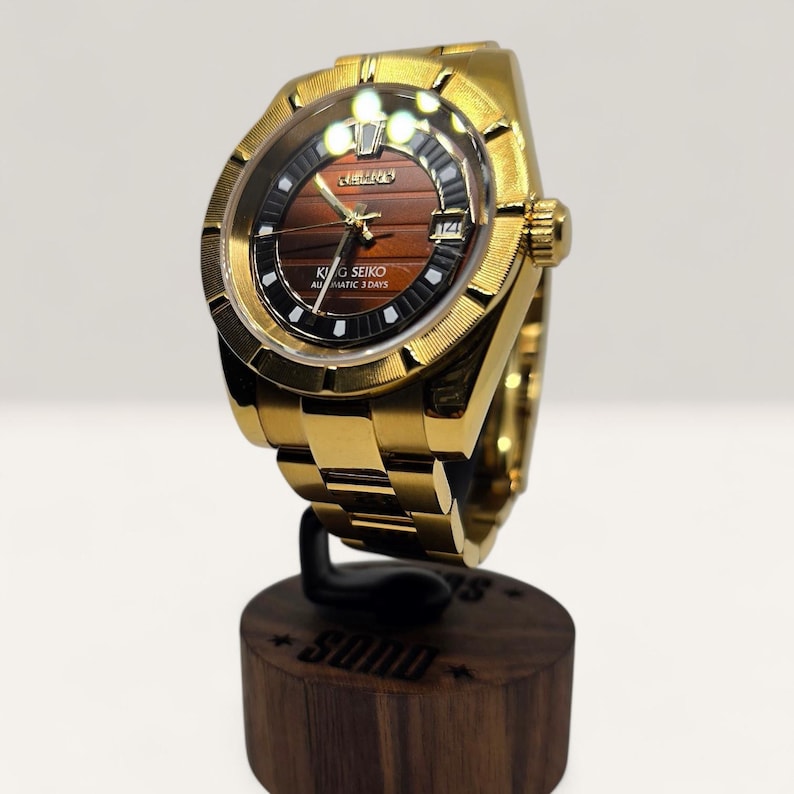 Op de afbeelding: Een goudkleurig polshorloge met een bruine wijzerplaat en een gouden band. Op de wijzerplaat staat de tekst "KING SEIKO AUTOMATIC 3 DAYS". Het horloge wordt gepresenteerd op een houten standaard.