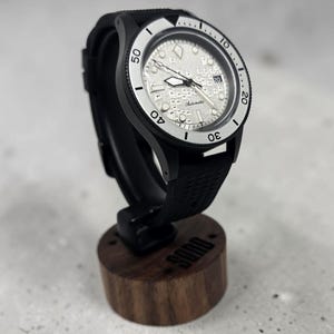 Puede incluir: Un reloj de pulsera automático con caja negra, esfera plateada texturizada con patrón de rejilla y correa de caucho negra. El bisel es plateado con números y marcadores negros. El reloj está montado sobre un soporte de madera cilíndrico.