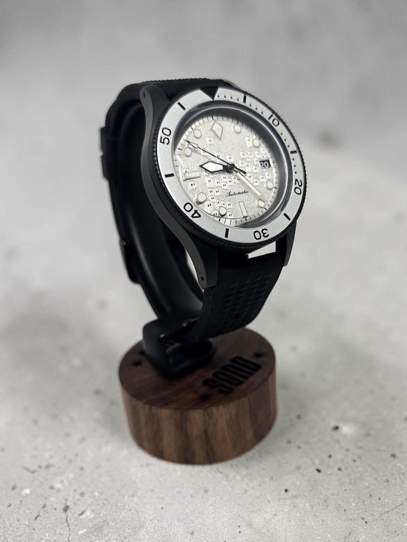 May include: An automatic wristwatch with a black case, a textured silver dial featuring a grid pattern, and a black rubber strap. The bezel is silver with black numerals and markers. The watch is mounted on a cylindrical wooden display stand.