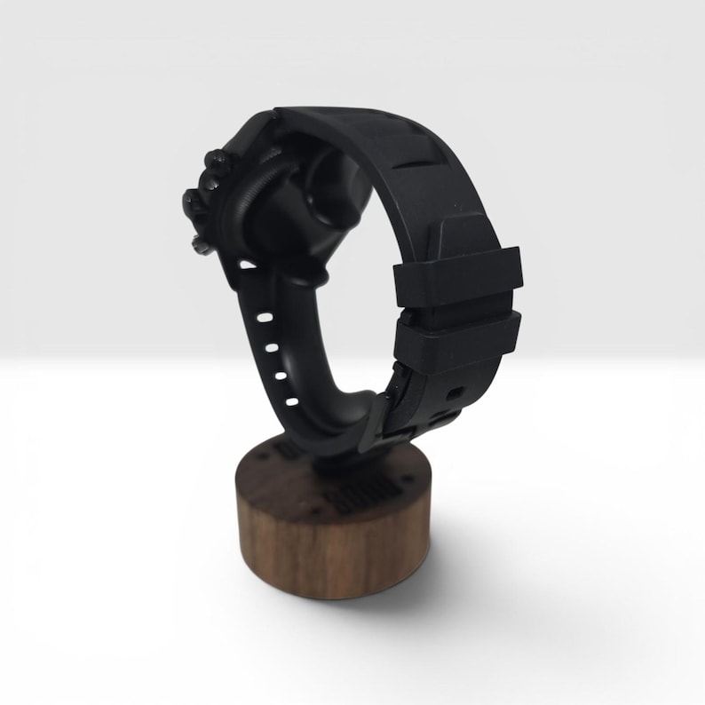 K&ouml;nnte beinhalten: Eine mattschwarze Armbanduhr mit strukturiertem Silikonarmband und Dornschlie&szlig;e, montiert auf einem zylindrischen, dunkelbraunen Holzst&auml;nder. Die Uhr verf&uuml;gt &uuml;ber ein rundes Geh&auml;use mit seitlichen Kn&ouml;pfen und eine durchgehend glatte, schwarze Oberfl&auml;che.