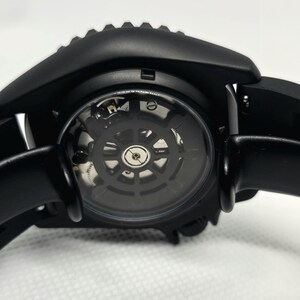 May include: A matte black wristwatch featuring an exhibition case back that reveals the intricate mechanical movement inside. The watch has a smooth black strap and a circular frame with a gear-like edge, showcasing the internal gears and springs.