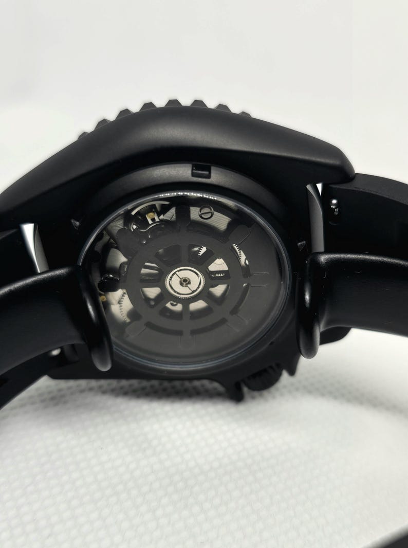 May include: A matte black wristwatch featuring an exhibition case back that reveals the intricate mechanical movement inside. The watch has a smooth black strap and a circular frame with a gear-like edge, showcasing the internal gears and springs.