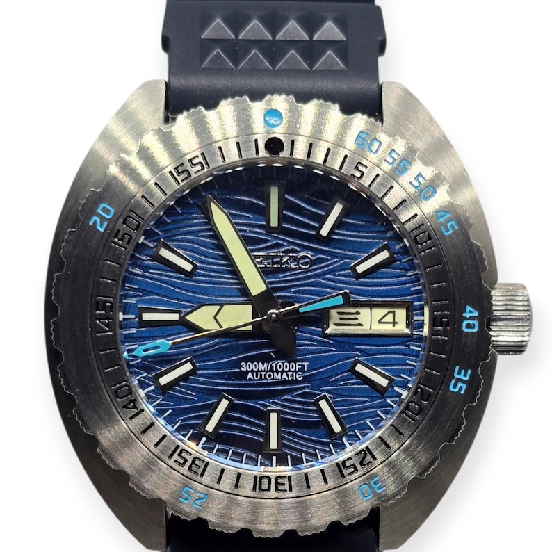 May include: A stainless steel wristwatch with a blue wave-patterned dial. The watch features luminous hands and markers, a date display, and a black rubber strap. The bezel has minute markers and a rotating design. The watch is water-resistant to 300 meters.