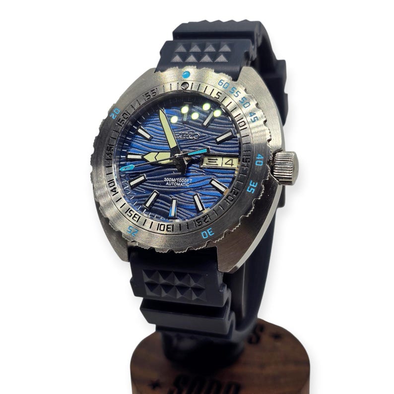 May include: A stainless steel dive watch with a blue wave-patterned dial and a black rubber strap. The watch features a rotating bezel with minute markers and the text "300M/1000FT AUTOMATIC". The date is displayed at the 3 o'clock position.