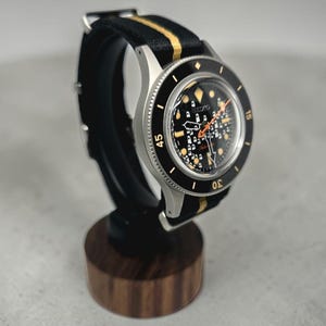 May include: A Seiko automatic wristwatch with a black dial featuring a grid pattern, luminous hour markers, and an orange second hand. The watch has a black rotating bezel with white numerals and is fitted with a black and gold striped nylon strap.