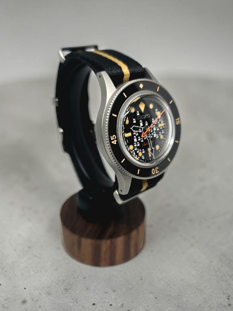 May include: A Seiko automatic wristwatch with a black dial featuring a grid pattern, luminous hour markers, and an orange second hand. The watch has a black rotating bezel with white numerals and is fitted with a black and gold striped nylon strap.