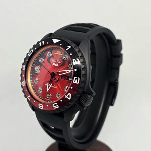 May include: A Seiko 5 Sports automatic wristwatch featuring a vibrant red dial with black graphic accents, a black rotating bezel with white numerals, and a black silicone strap. The watch face displays the Seiko logo and the word Automatic.