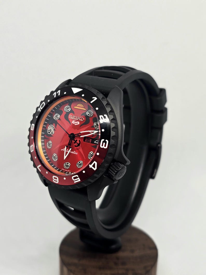 May include: A Seiko 5 Sports automatic wristwatch featuring a vibrant red dial with black graphic accents, a black rotating bezel with white numerals, and a black silicone strap. The watch face displays the Seiko logo and the word Automatic.