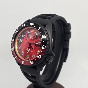 May include: A black Seiko 5 automatic watch with a red dial featuring a straw hat icon at the twelve o'clock position and fist symbols as hour markers. The watch has a black rotating bezel with white numerals and a black silicone rubber strap.