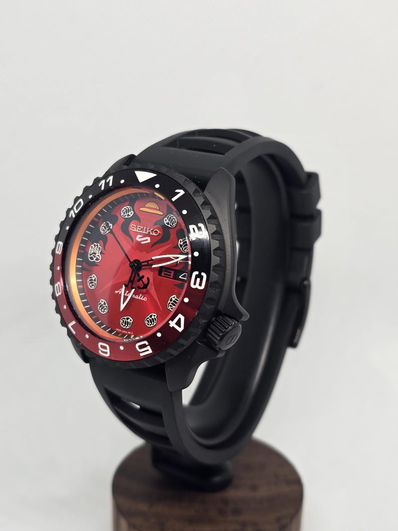 May include: A black Seiko 5 automatic watch with a red dial featuring a straw hat icon at the twelve o'clock position and fist symbols as hour markers. The watch has a black rotating bezel with white numerals and a black silicone rubber strap.