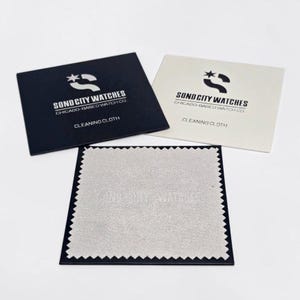 Puede incluir: Dos sobres rectangulares para pa&ntilde;os de limpieza, uno negro y otro blanco, impresos con el logotipo y el texto Sond City Watches Chicago-Based Watch Co. Debajo hay un pa&ntilde;o de limpieza cuadrado de color gris claro con bordes dentados y relieve.