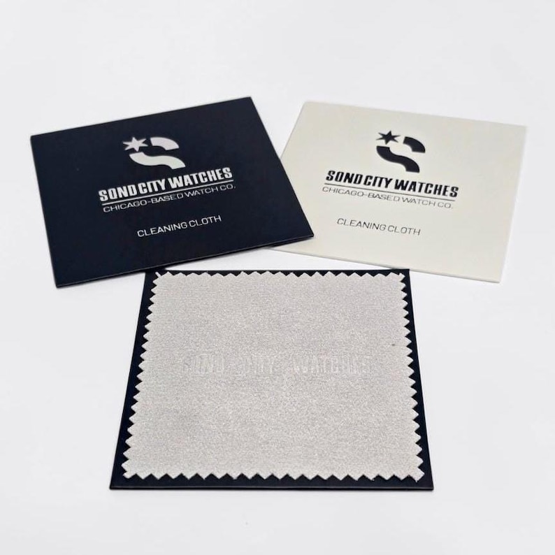 Puede incluir: Dos sobres rectangulares para pa&ntilde;os de limpieza, uno negro y otro blanco, impresos con el logotipo y el texto Sond City Watches Chicago-Based Watch Co. Debajo hay un pa&ntilde;o de limpieza cuadrado de color gris claro con bordes dentados y relieve.