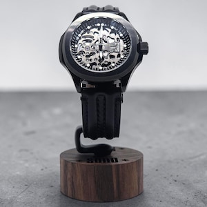 May include: A black wristwatch with a skeleton dial revealing internal mechanical components, featuring a black rubber strap. The watch is mounted on a cylindrical wooden display stand with the word "SUND" engraved on the front side.