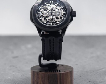 May include: A black wristwatch with a skeleton dial revealing internal mechanical components, featuring a black rubber strap. The watch is mounted on a cylindrical wooden display stand with the word "SUND" engraved on the front side.
