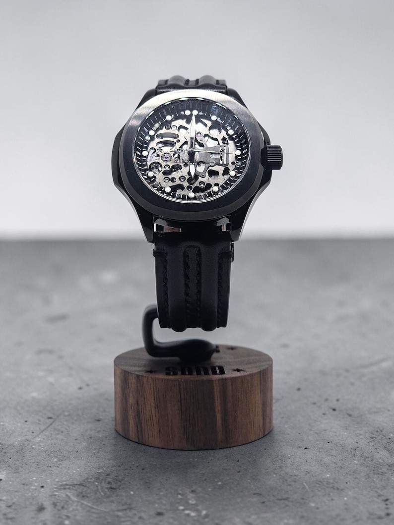 May include: A black wristwatch with a skeleton dial revealing internal mechanical components, featuring a black rubber strap. The watch is mounted on a cylindrical wooden display stand with the word "SUND" engraved on the front side.