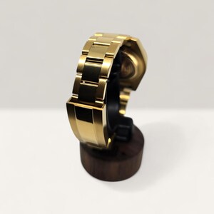 Op de afbeelding: Een goudkleurig polshorloge met bijpassende band wordt gepresenteerd op een houten horlogestandaard. Het horloge heeft een klassiek ontwerp met een rechthoekige wijzerplaat en een band in schakelstijl. Het horloge wordt gepresenteerd tegen een witte achtergrond.