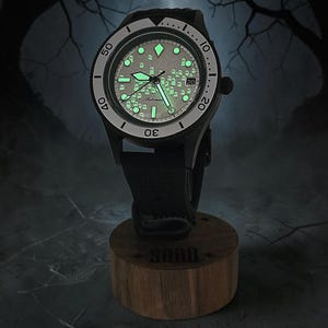 May include: A black automatic wristwatch with a glow-in-the-dark dial featuring a pattern of small ghost figures. The watch has a rotating bezel with numerical markers, luminous hands, and a date window, mounted on a cylindrical wooden stand.