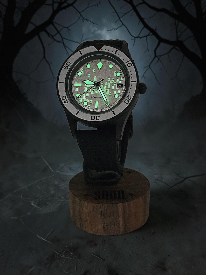 May include: A black automatic wristwatch with a glow-in-the-dark dial featuring a pattern of small ghost figures. The watch has a rotating bezel with numerical markers, luminous hands, and a date window, mounted on a cylindrical wooden stand.