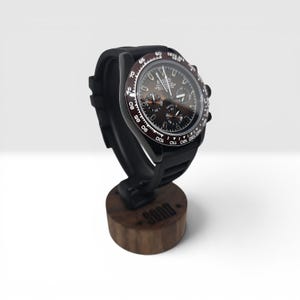 K&ouml;nnte beinhalten: Eine schwarze Chronographen-Armbanduhr mit dunkelbraunem Zifferblatt und Tachymeter-L&uuml;nette, die den Schriftzug Seiko und Daytona auf dem Zifferblatt tr&auml;gt. Die Uhr ist auf einem dunkelbraunen Holzst&auml;nder mit dem Wort SOOD auf der Vorderseite montiert.