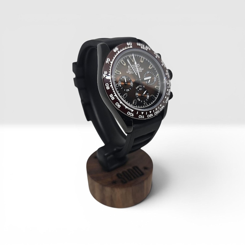K&ouml;nnte beinhalten: Eine schwarze Chronographen-Armbanduhr mit dunkelbraunem Zifferblatt und Tachymeter-L&uuml;nette, die den Schriftzug Seiko und Daytona auf dem Zifferblatt tr&auml;gt. Die Uhr ist auf einem dunkelbraunen Holzst&auml;nder mit dem Wort SOOD auf der Vorderseite montiert.
