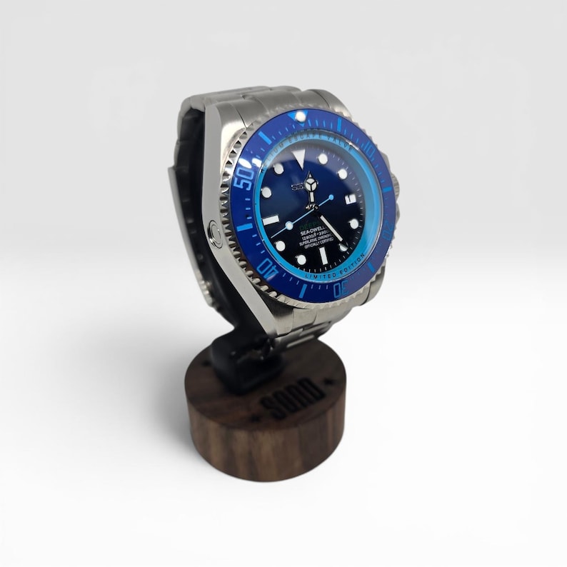 May include: A stainless steel wristwatch with a blue and black rotating bezel, a dark blue dial featuring white hour markers, and the text SEA-DWELLER 12800ft/3900m LIMITED EDITION. The watch is mounted on a cylindrical dark wood display stand.