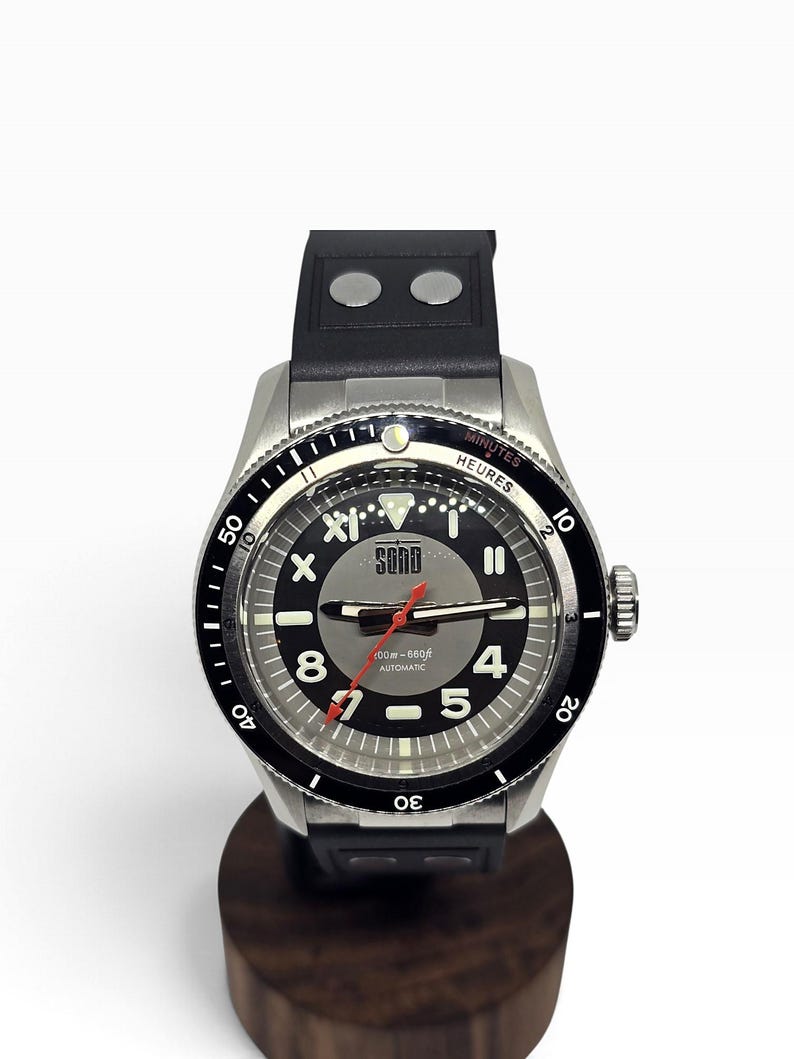 Puede incluir: Un reloj autom&aacute;tico de acero inoxidable con esfera negra, que presenta el nombre de la marca SOND, n&uacute;meros ar&aacute;bigos blancos 4, 5, 7, 8 y 11, un segundero rojo y un bisel giratorio negro con marcas blancas. Correa de caucho negra con dos remaches plateados.