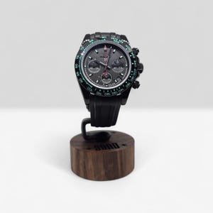 May include: A black chronograph watch with a dark gray dial, three sub-dials, and a tachymeter bezel with green numerals. The watch features a black silicone strap and is mounted on a cylindrical dark wood stand with the word SONO engraved.