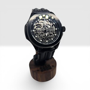 May include: A black skeleton watch with a round case, a black leather strap, and a visible mechanical movement. The watch face features white hour markers and silver hands. It is mounted on a cylindrical wooden stand with the word SORD engraved.