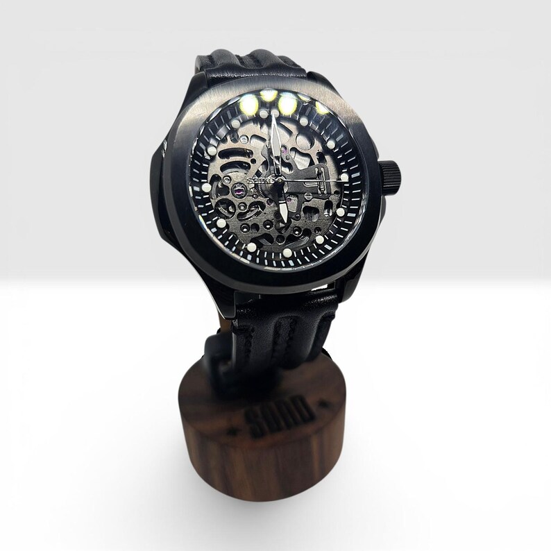 May include: A black skeleton watch with a round case, a black leather strap, and a visible mechanical movement. The watch face features white hour markers and silver hands. It is mounted on a cylindrical wooden stand with the word SORD engraved.