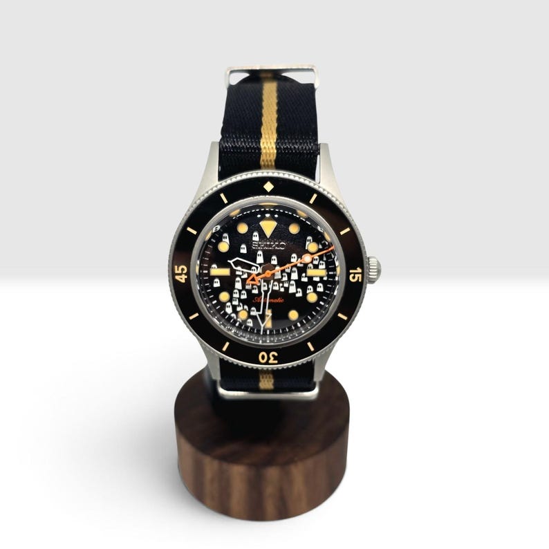 May include: An automatic wristwatch with a silver-tone case and a black dial featuring a repeating white ghost pattern. The watch has a black nylon strap with a central gold stripe, a rotating black bezel with gold numerals, and orange hands.