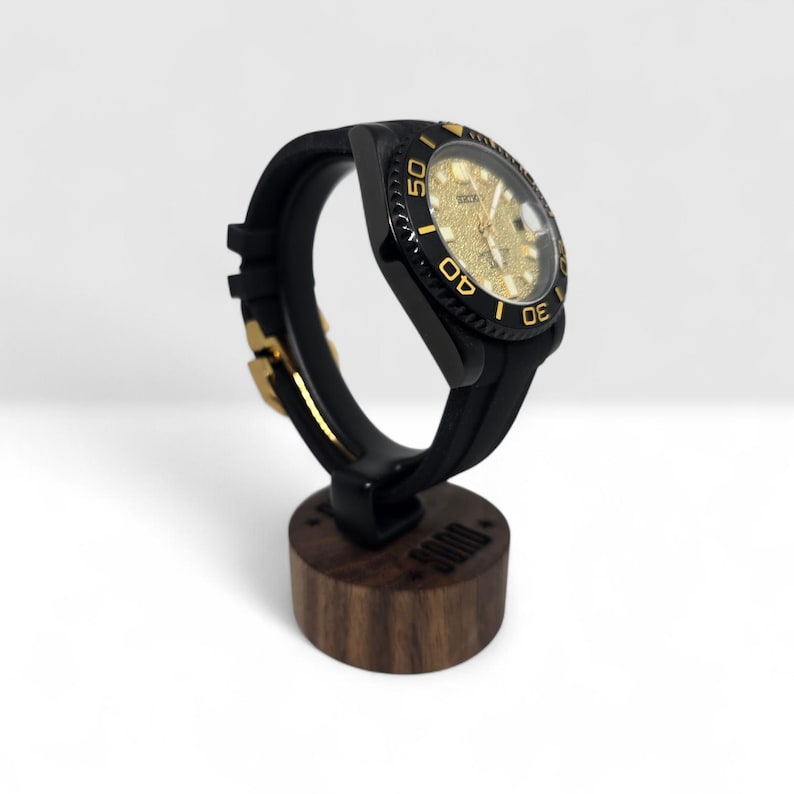 Pu&ograve; includere: Un orologio da polso nero con quadrante dorato testurizzato e lunetta nera con numeri dorati. L'orologio presenta un cinturino in silicone nero con fibbia dorata ed &egrave; montato su un supporto cilindrico in legno scuro con la scritta SOND.