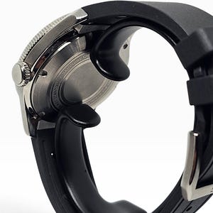 Puede incluir: Un reloj de pulsera con correa de caucho negra y caja de acero inoxidable plateado. Se observa la parte posterior de la caja, con detalles de metal cepillado circular y el texto SAPPHIRE CRYSTAL y WATER RESISTANT 200M.